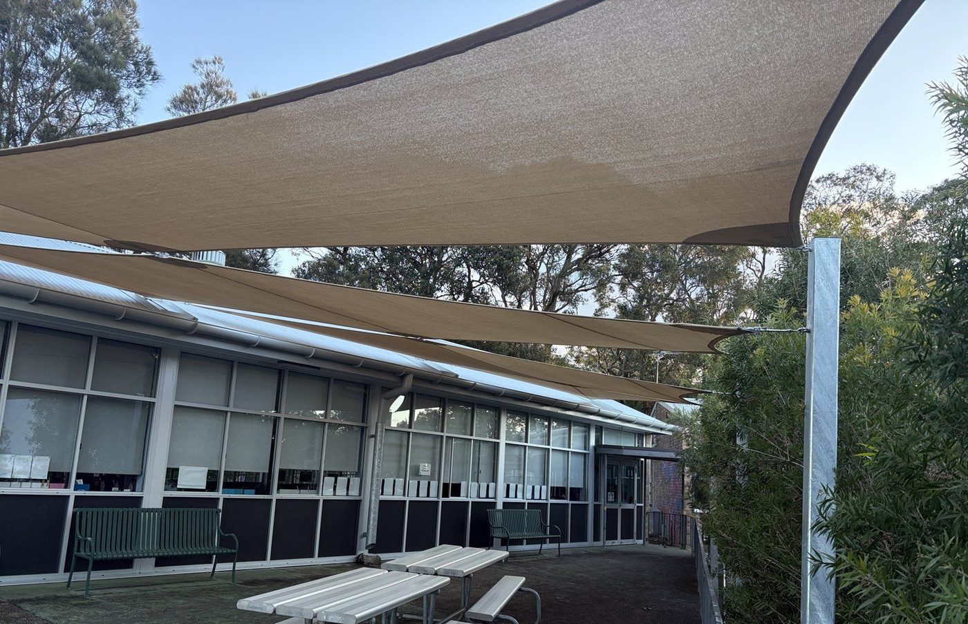 Sand shade sails angled over a paved courtyard with picnic tables beside a low building., Mona Vale