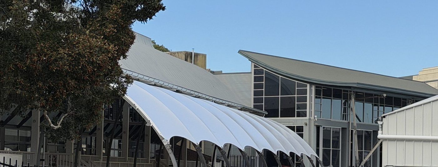 A white curved membrane canopy on steel arches beside a large glass-fronted building., Moore Park