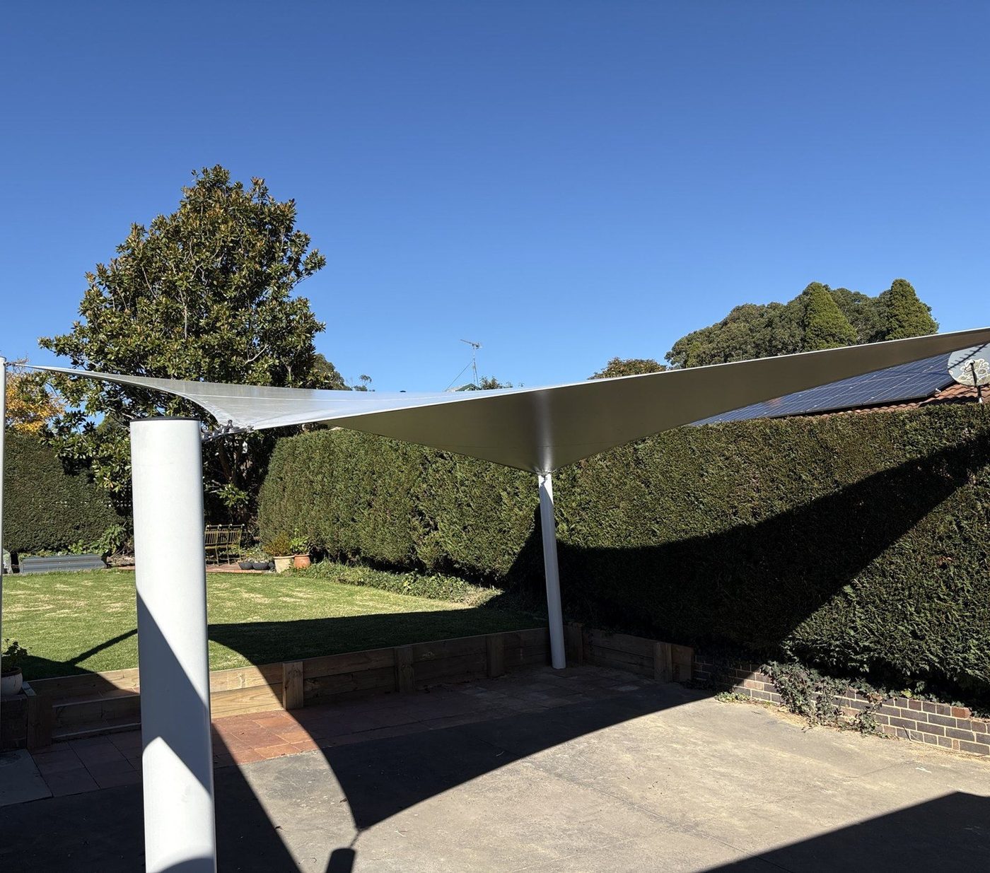Sand-coloured shade sail on white steel posts over a paved courtyard beside a tall clipped hedge., Moss Vale