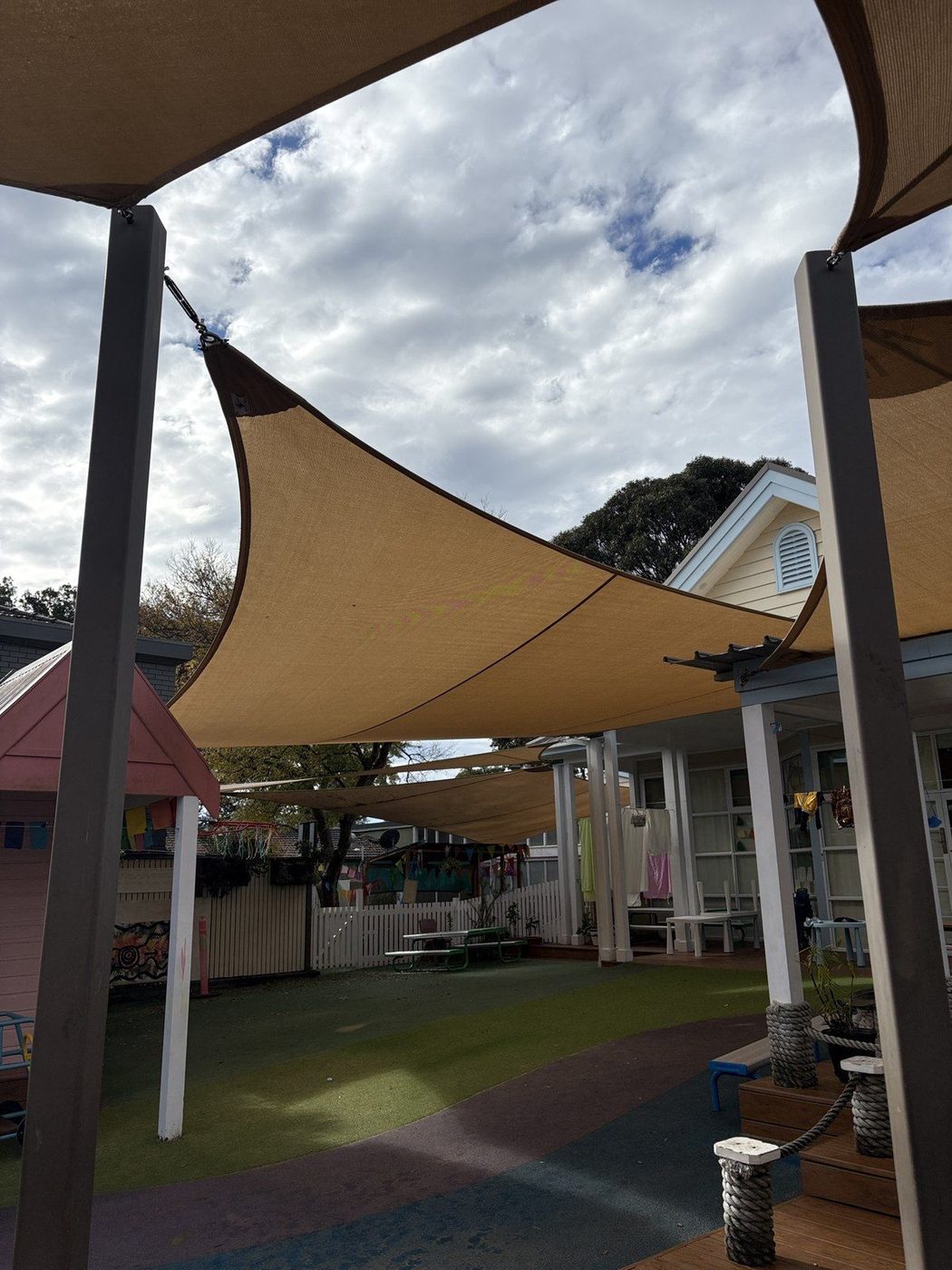 Sand shade sails on steel posts above a synthetic turf playground with a cubby house and picket fence., Narrabeen