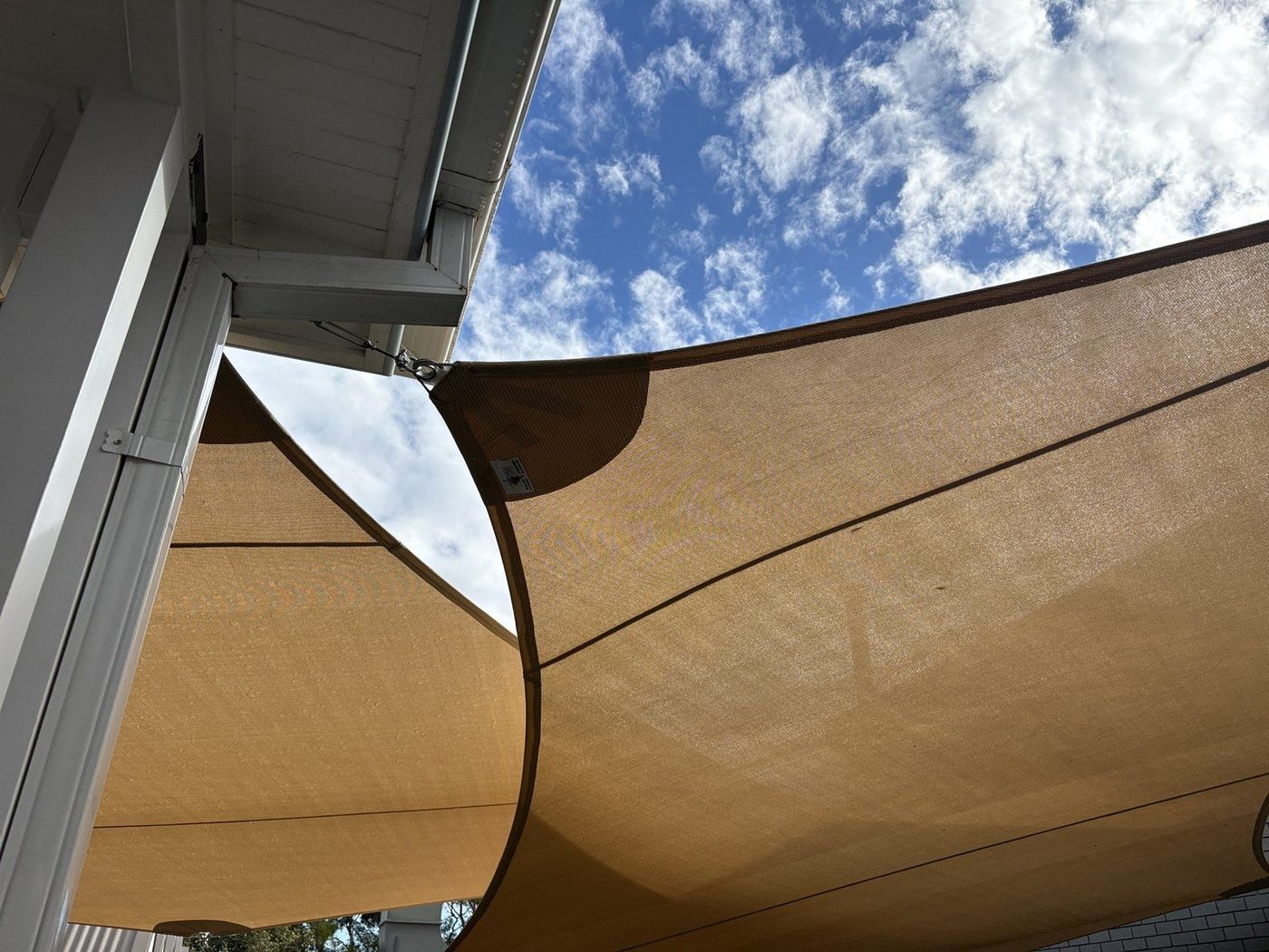 Two sand-coloured shade sails seen from below against a blue sky beside the white eave of a building., Narrabeen