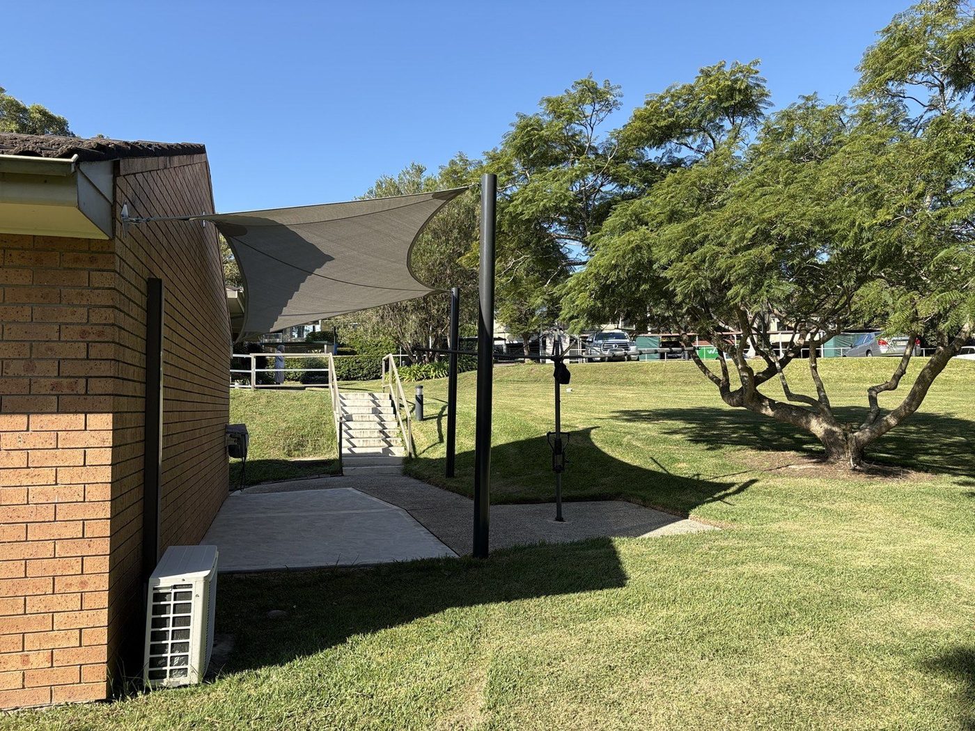 Grey shade sail from a brick wall to black posts above a paved path and stairs on a lawn., Narrabeen