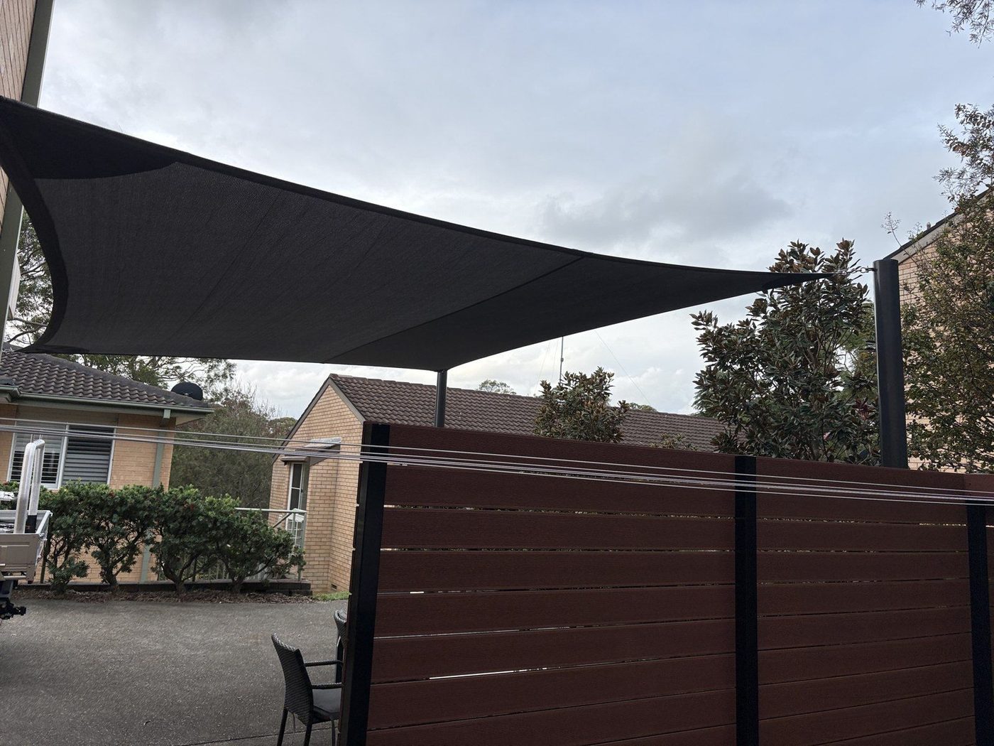 Charcoal shade sails above a paved common area beside a dark timber privacy screen., Narrabeen