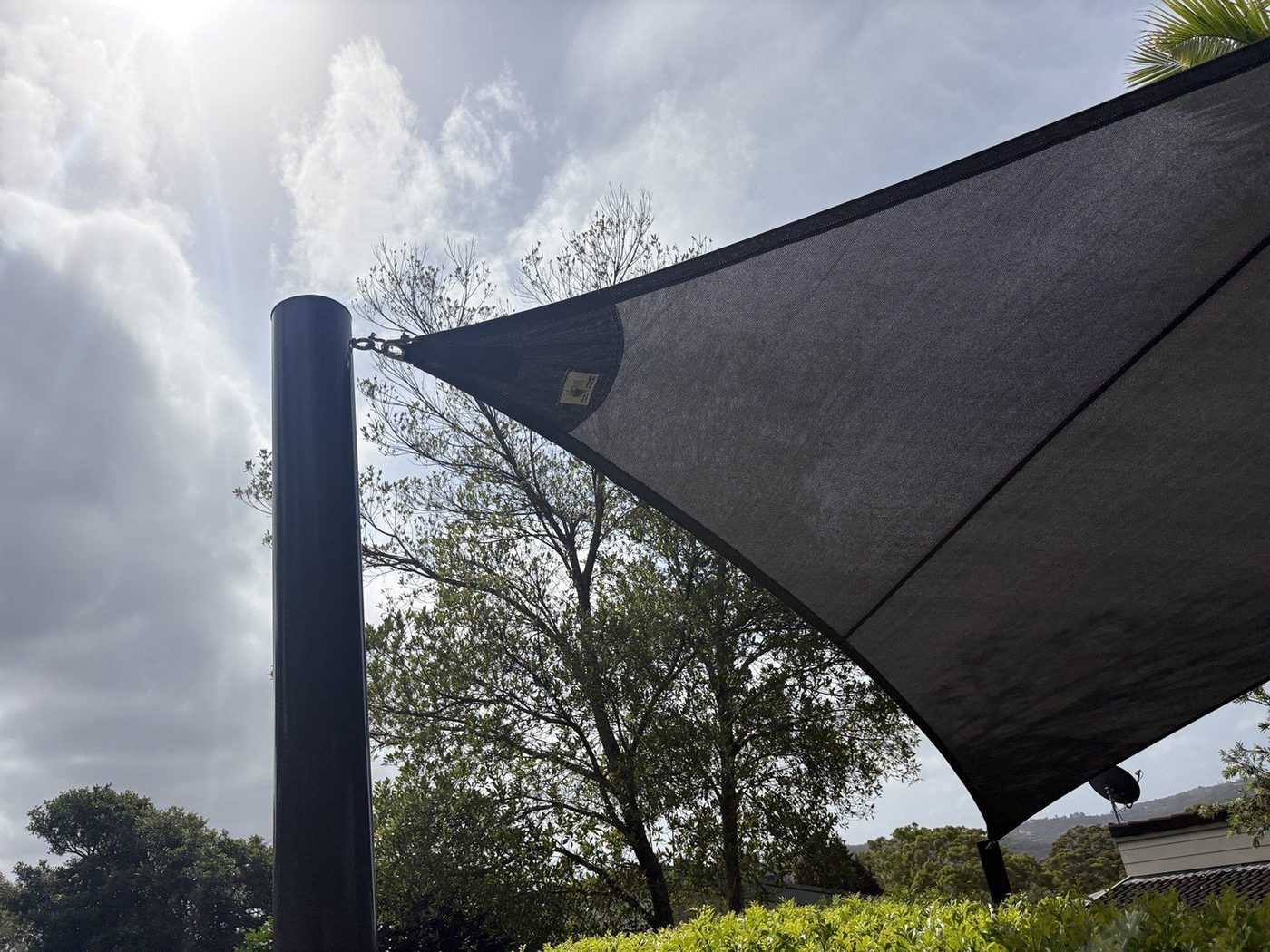 A charcoal shade sail curving from a dark steel post against a bright, cloud-streaked sky., Narrabeen