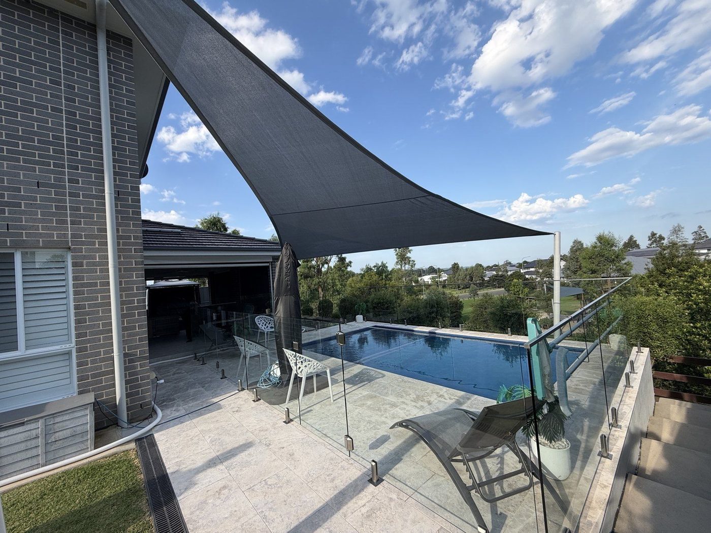 A charcoal shade sail angled over a tiled pool terrace with glass pool fencing., North Richmond