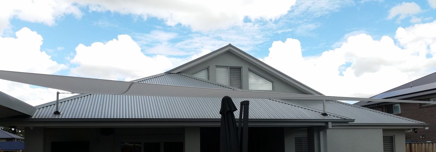 Two white shade sails stretched above the metal roofline of a single-storey home under a cloudy sky., North Rocks