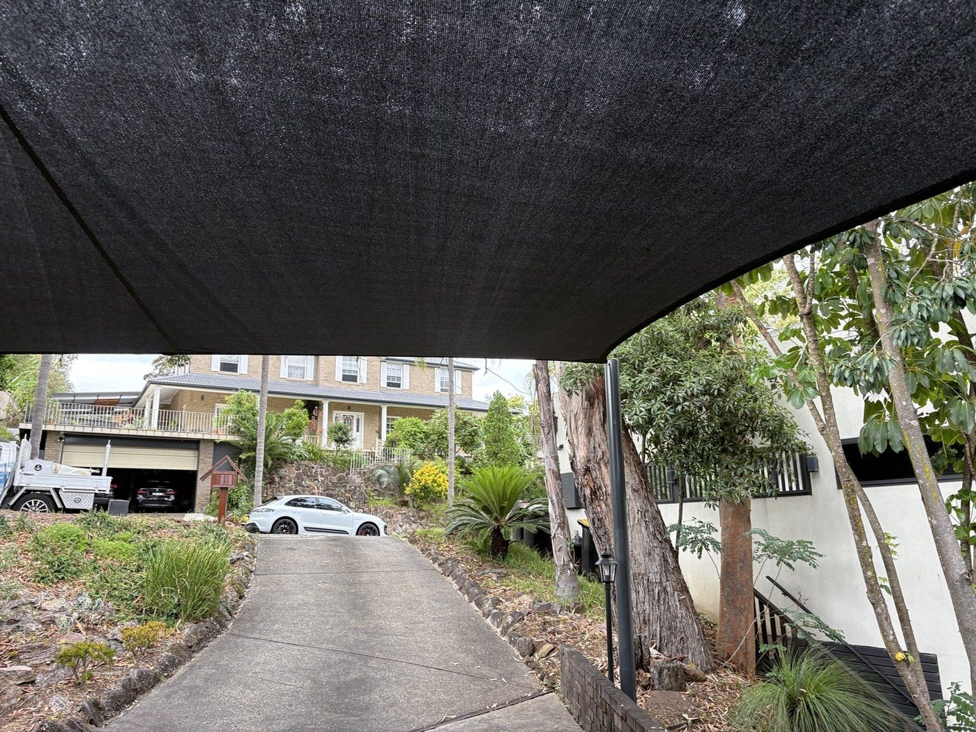 Black shade sail seen from beneath, spanning a sloping driveway beside a stone retaining wall, North Rocks