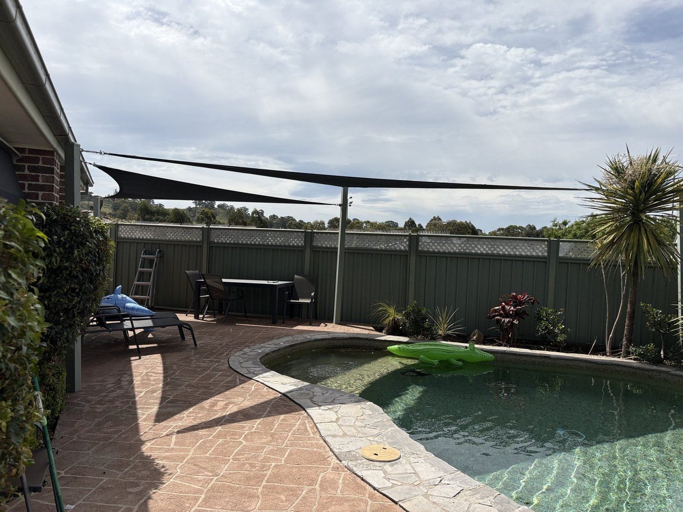 A charcoal shade sail over a backyard pool with a paved surround under grey cloud., Point Clare