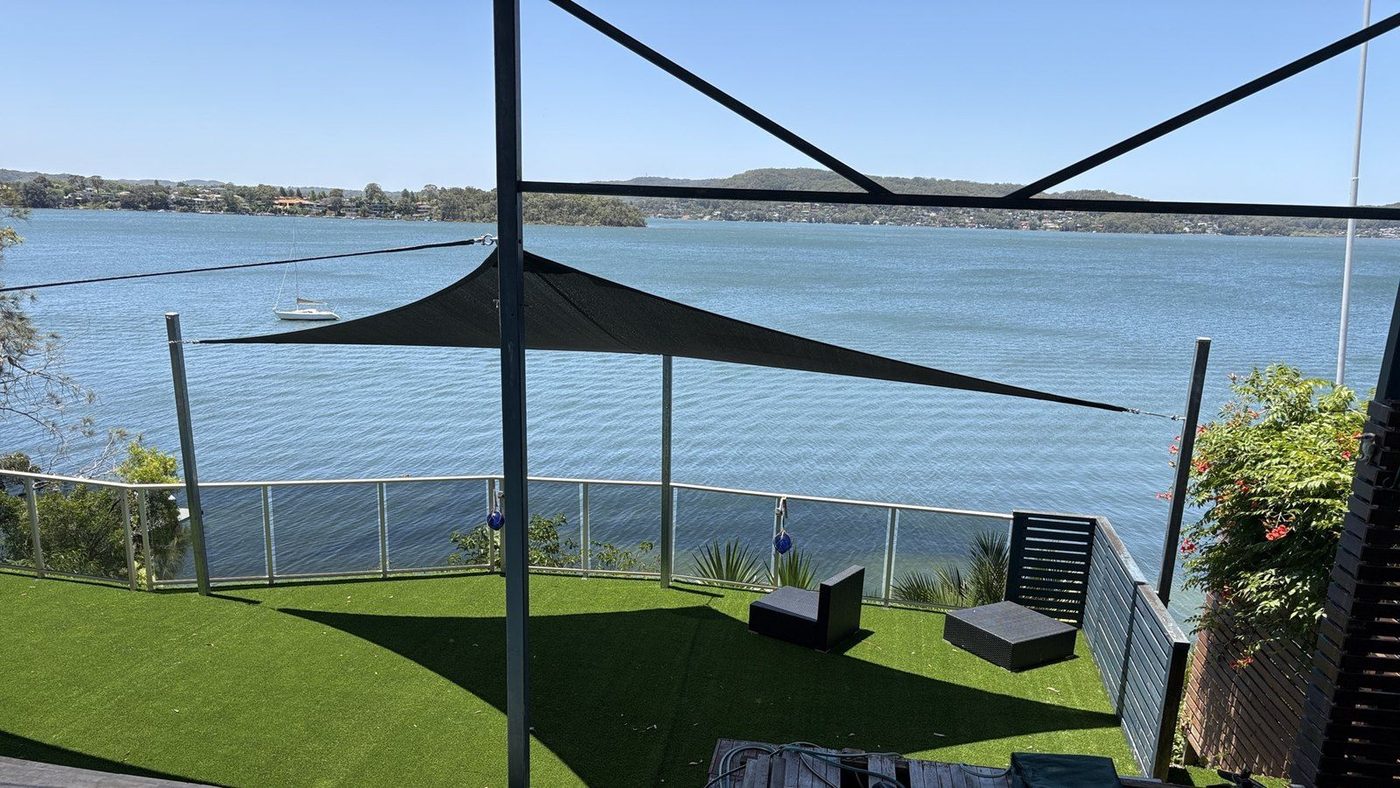 A black shade sail on steel posts over a turf lawn behind a glass balustrade with a waterway beyond., Point Clare