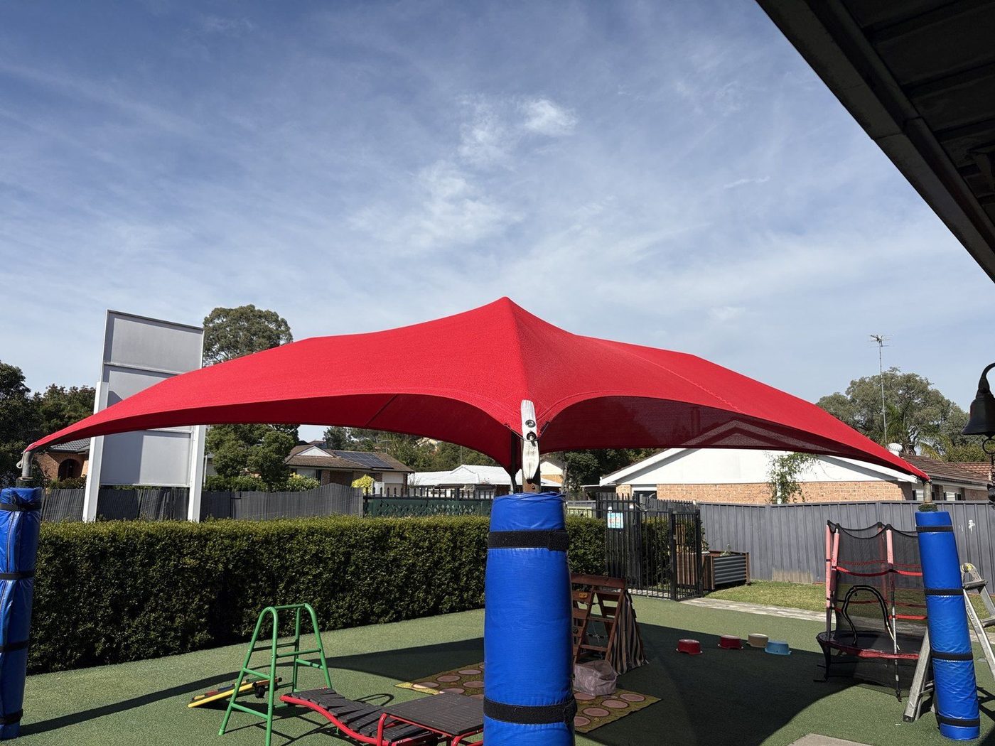 Red shade sail peaked on a central padded post above a play area with a hedge and timber fence., Raby