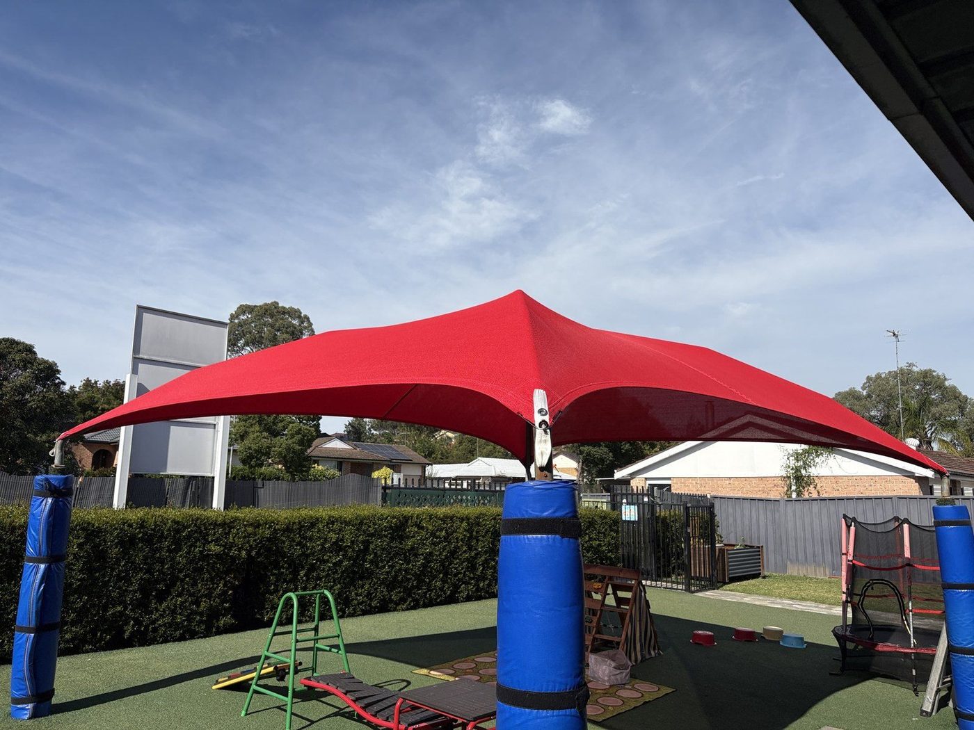 Red shade sail on a centre post above a playground with padded posts and green soft fall., Raby