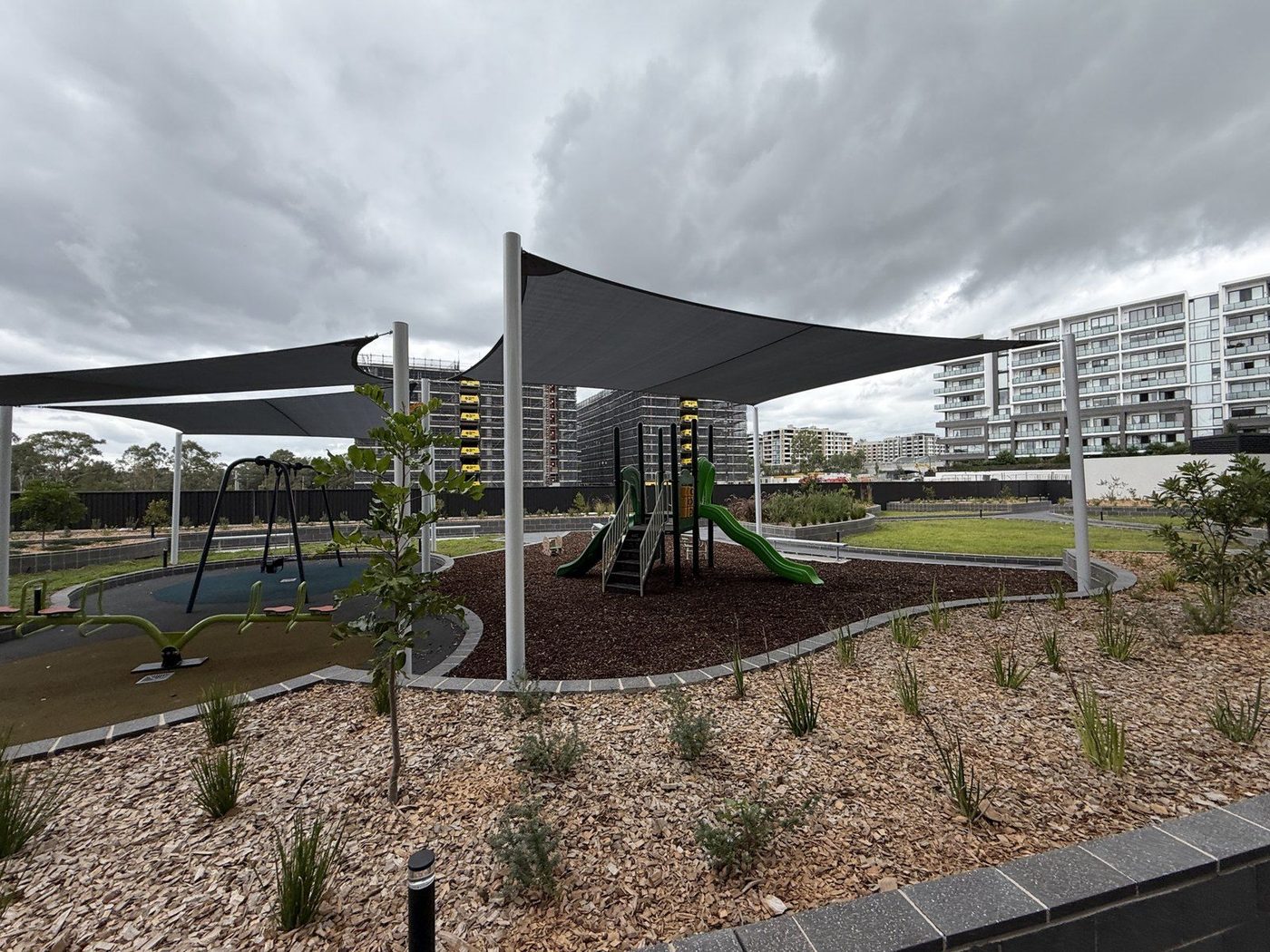 Charcoal shade sails on white posts above a playground in an apartment complex courtyard., Rouse Hill