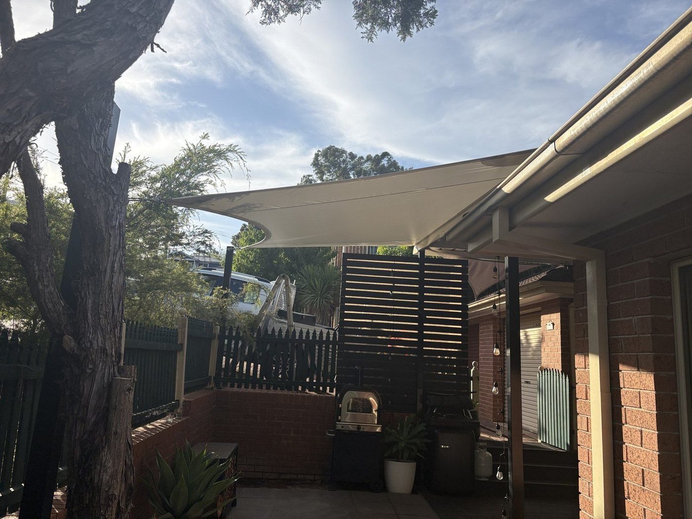 Sand shade sail laced to the gutter above a paved back patio with a barbecue and festoon lights., Ryde