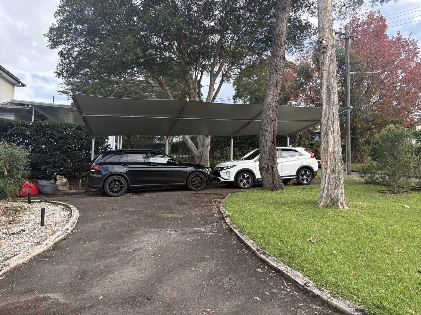 A dark cantilever shade sail over parked cars beside a lawn and mature trees., Saint Ives