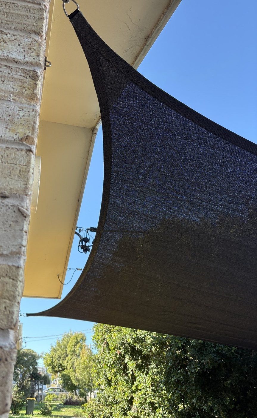 A charcoal shade sail anchored to the eave of a brick home, seen from below against blue sky., South Wentworthville