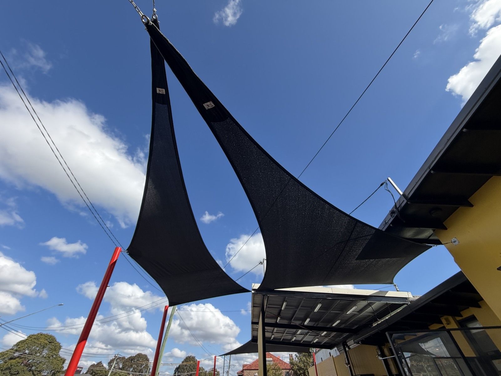 Two black mesh shade sails viewed from below against blue sky, with red posts and visible tension fittings.
