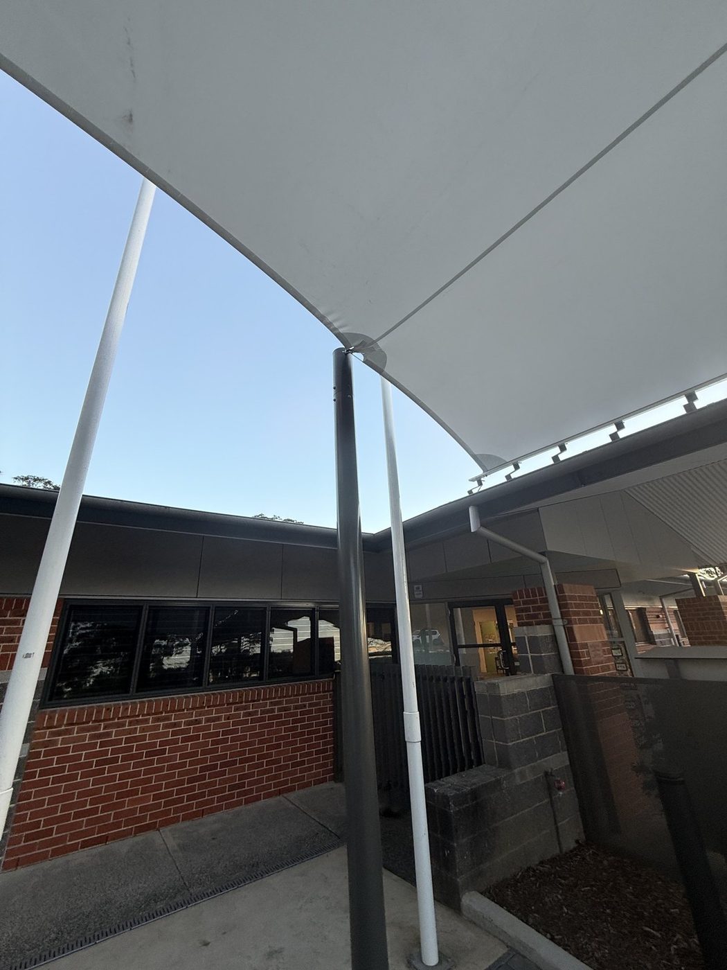 A white waterproof membrane canopy over a paved entry courtyard beside a low brick building., West Gosford