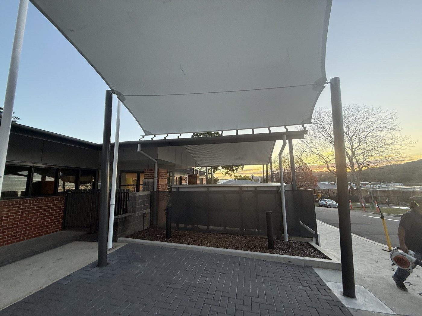 A white shade sail over a brick-paved entry at dusk, with a fenced road and hills beyond., West Gosford