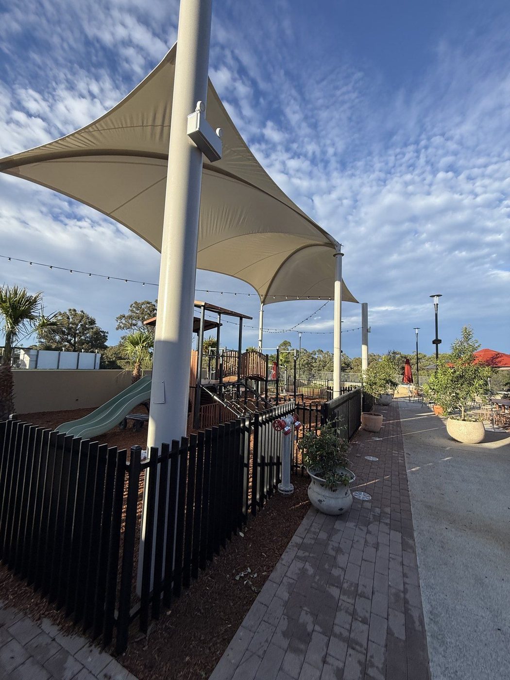 Cream conic shade sails on white steel posts above a fenced playground beside a paved courtyard., Wilton