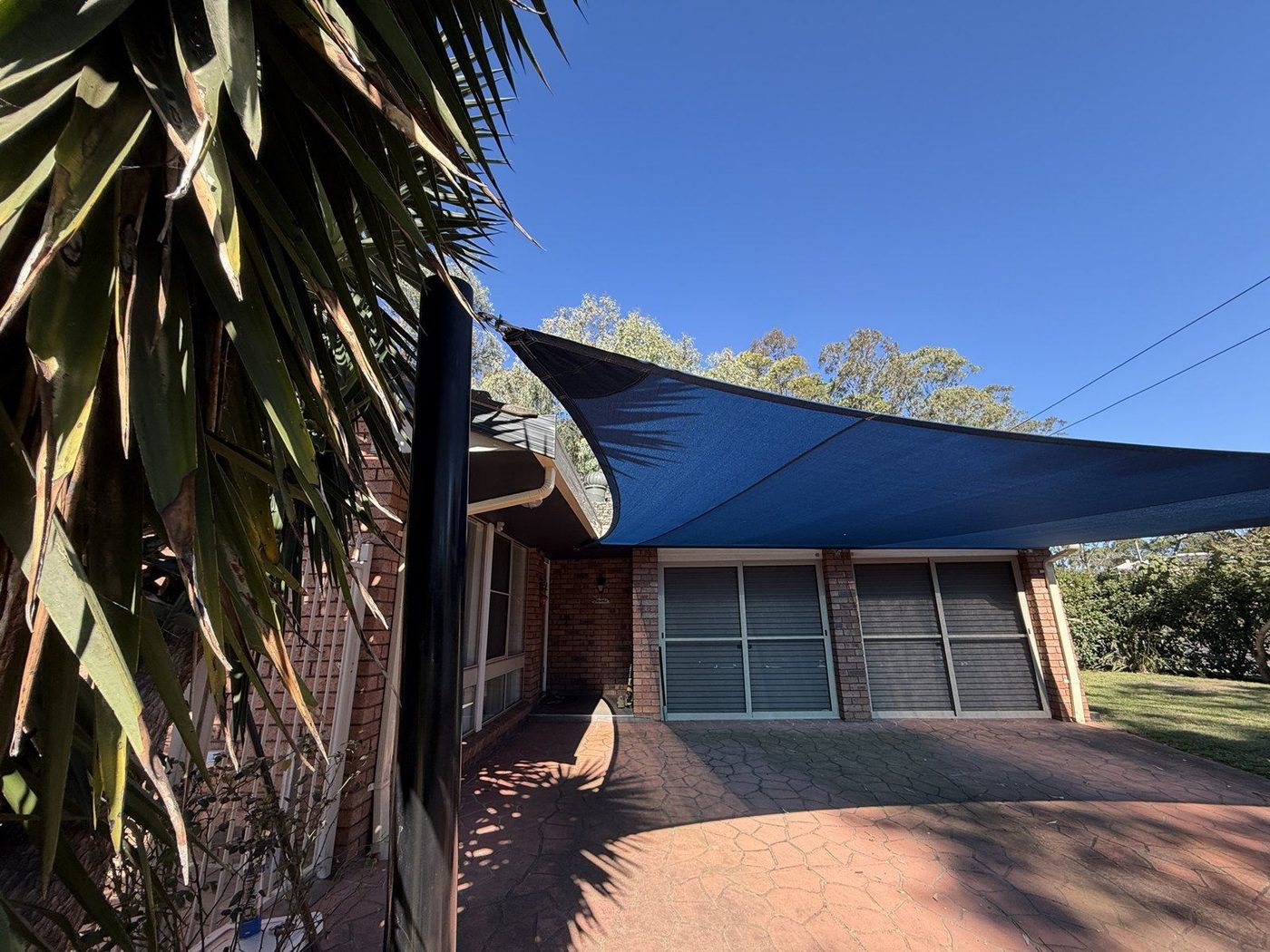 A blue shade sail over a paved driveway beside a brick home under clear sky., Wilton