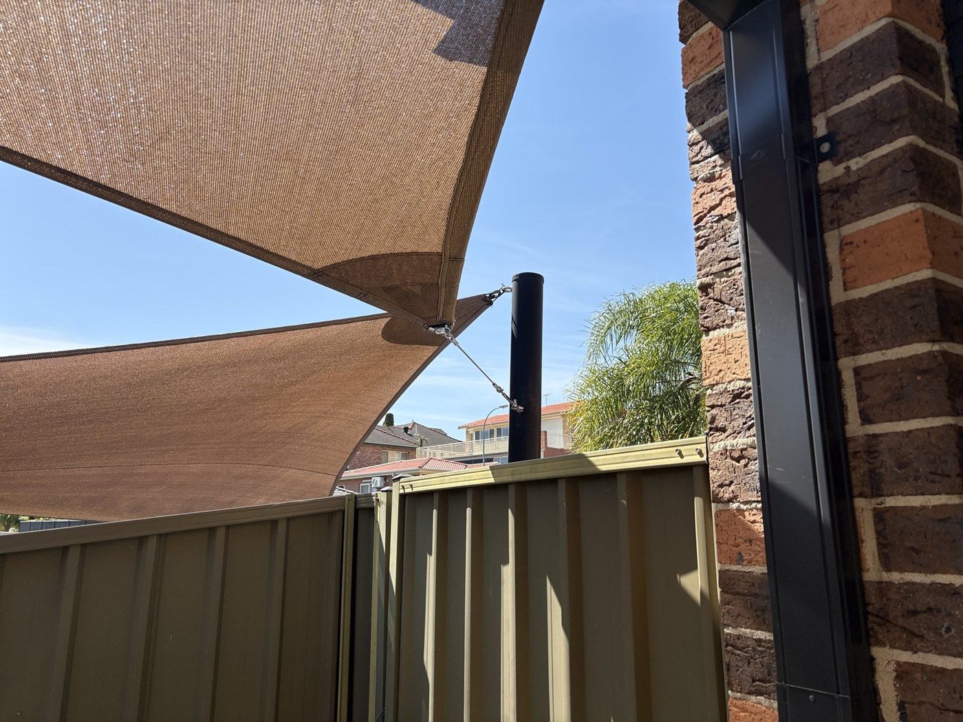 A sand shade sail corner tensioned by a cable and turnbuckle to a black steel post., Woodbine