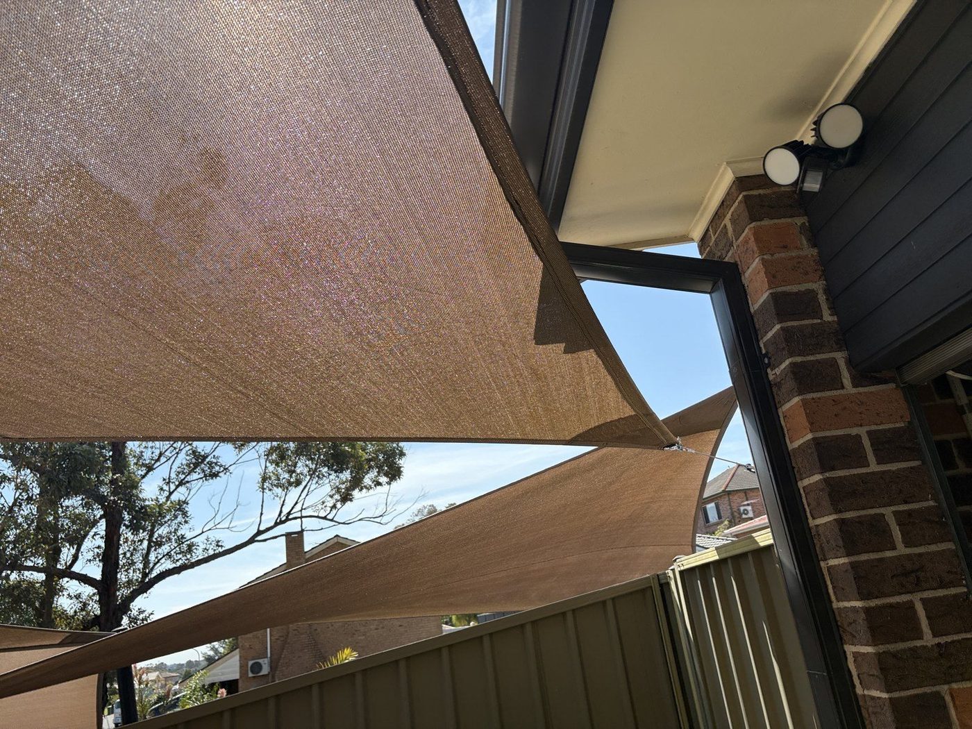 Overlapping sand shade sails seen from beneath a brick patio eave against blue sky., Woodbine