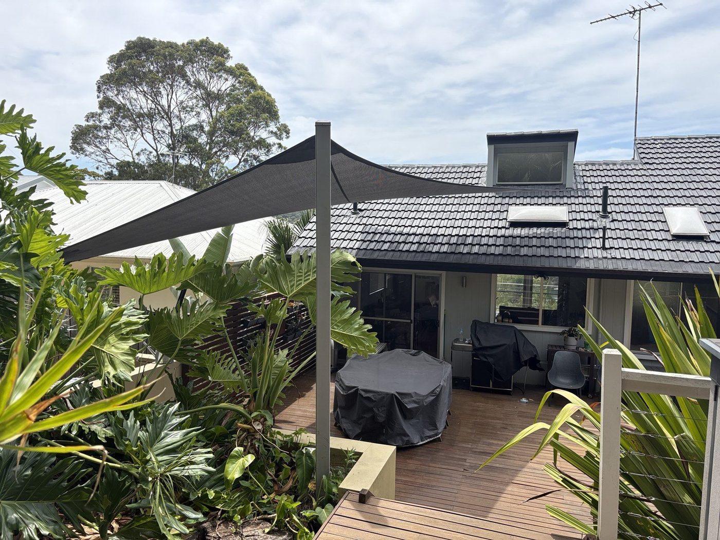 A charcoal shade sail on a steel post shading a timber deck beside a tiled-roof home., Woronora